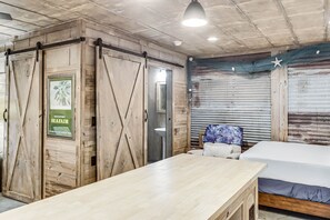 A room with a wooden table, a bed, a bathroom with a sink, and a door with a poster advertising a seafair.