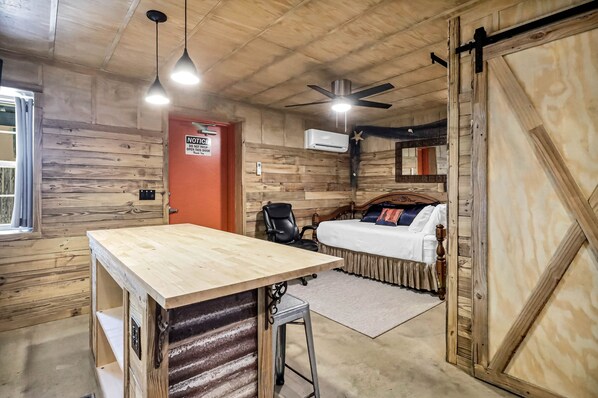 Quarto rústico com teto de madeira, cama com colcha branca, escrivaninha com cadeira e ventilador de teto.