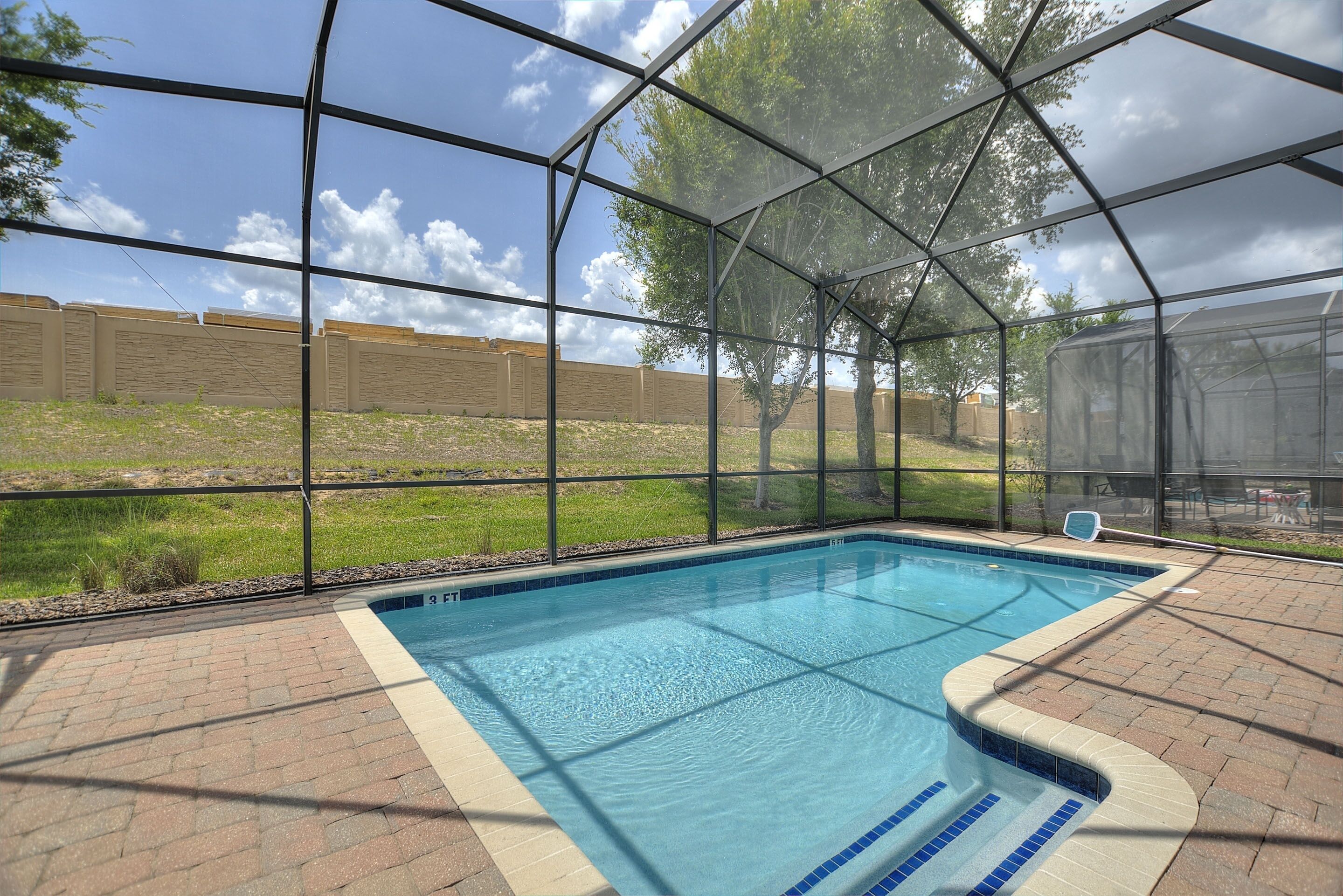 A swimming pool with a clear view of the surrounding area, including a brick patio and a grassy field.