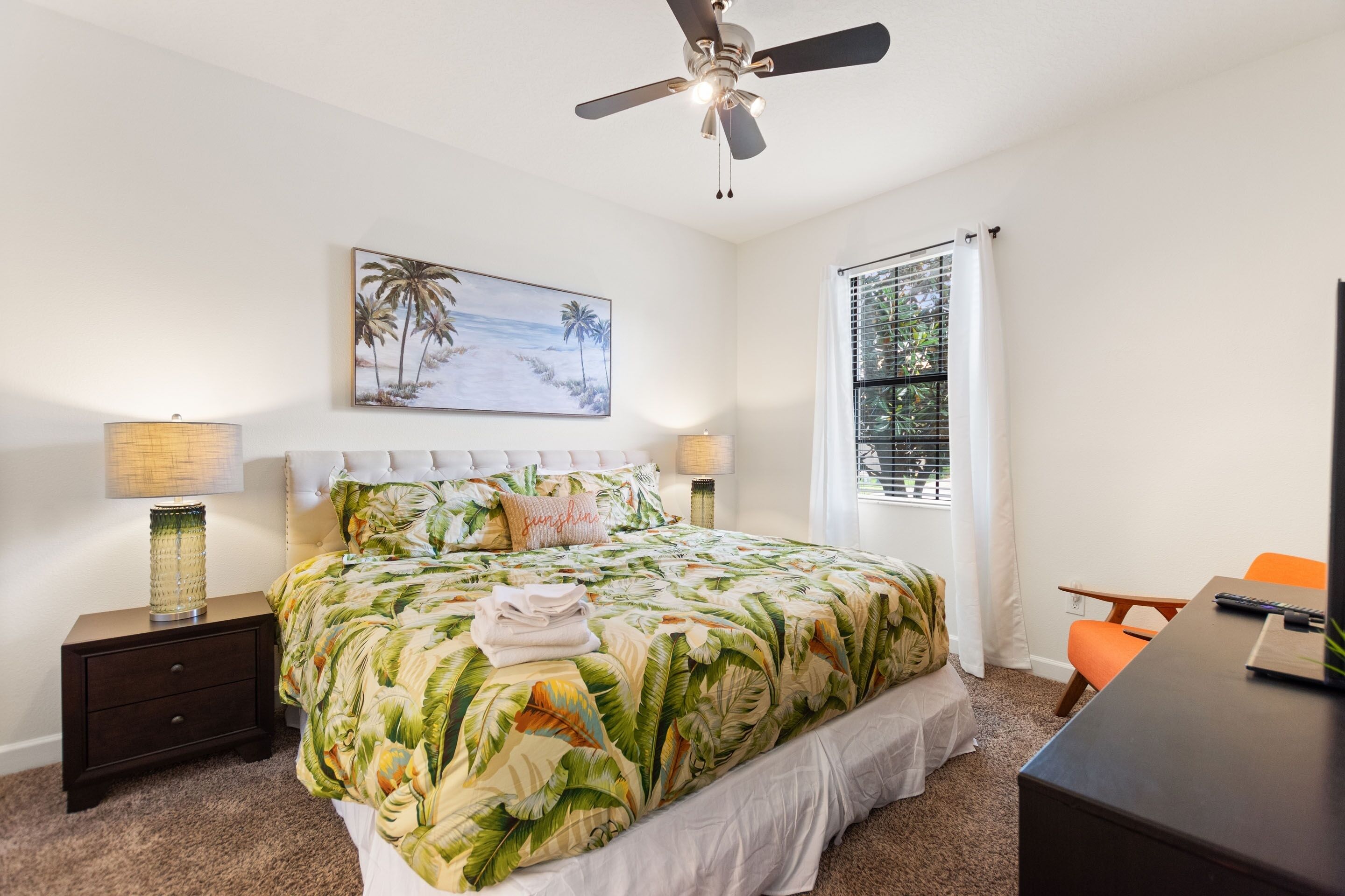 A bedroom with a bed, nightstands, a desk, a chair, a ceiling fan, and a window with blinds.