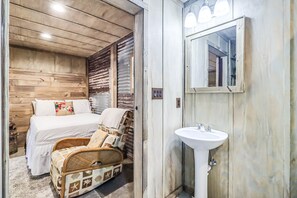 Un dormitorio con techo de madera, una cama con ropa de cama blanca, una silla de mimbre y un lavamanos blanco con espejo.