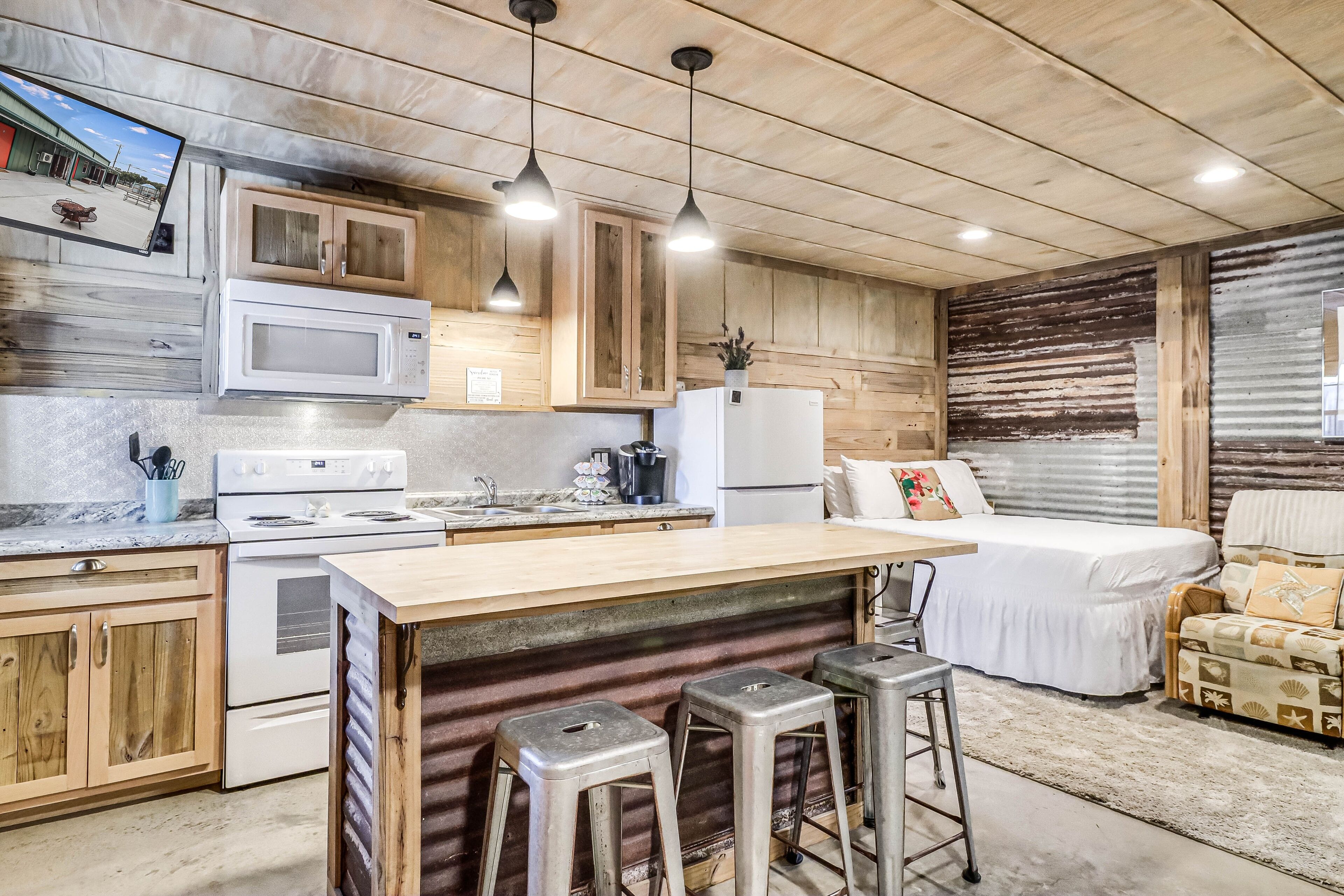 A kitchen with wooden cabinets, a microwave, a stove, and a refrigerator. There are stools and a bed in the room.