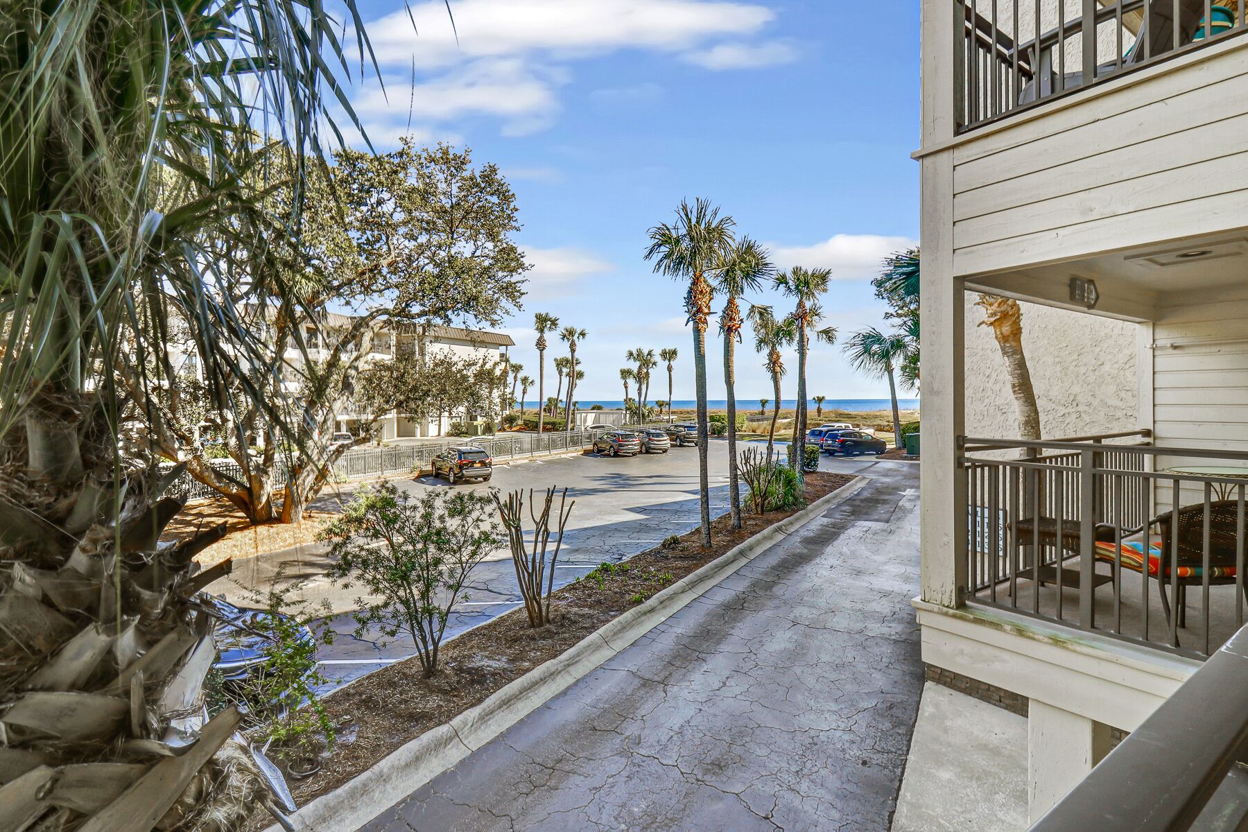 A street with palm trees, a parking lot, and a building with a balcony.