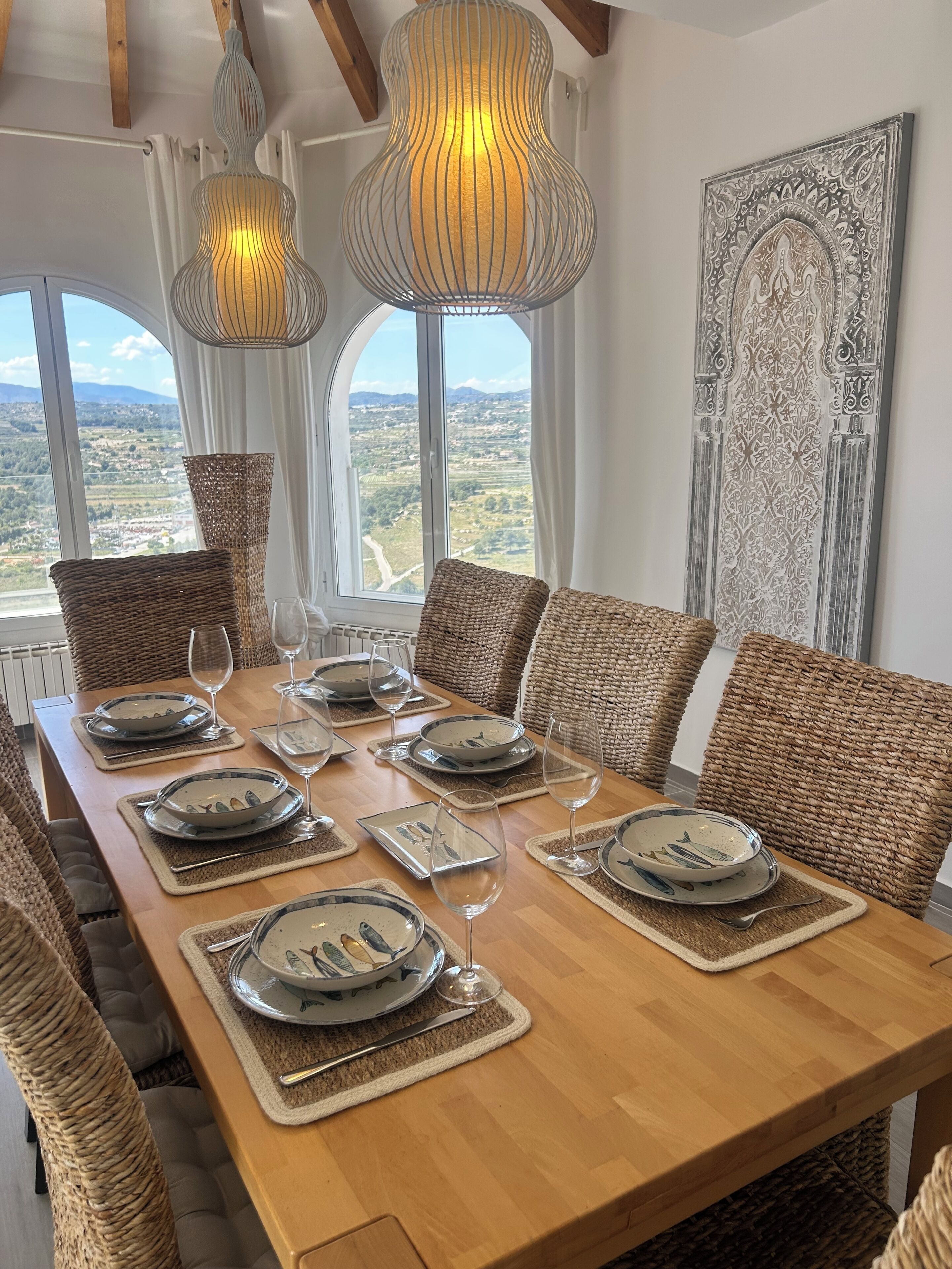 A dining table set with plates, glasses, and cutlery, surrounded by woven chairs, with a view of the countryside through large windows.