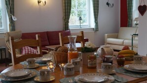 A breakfast table set with plates, cups, and a teapot, surrounded by chairs and a sofa in a cozy room with a window and curtains.