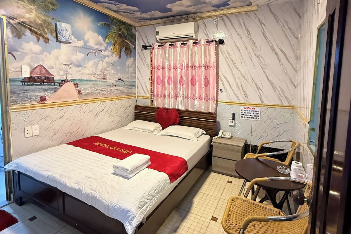 A hotel room with a bed, a small table, a chair, a ceiling fan, and a mural of a tropical scene.