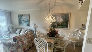 A living room with a floral sofa, a dining table with chairs, a hanging light fixture, and a painting on the wall.