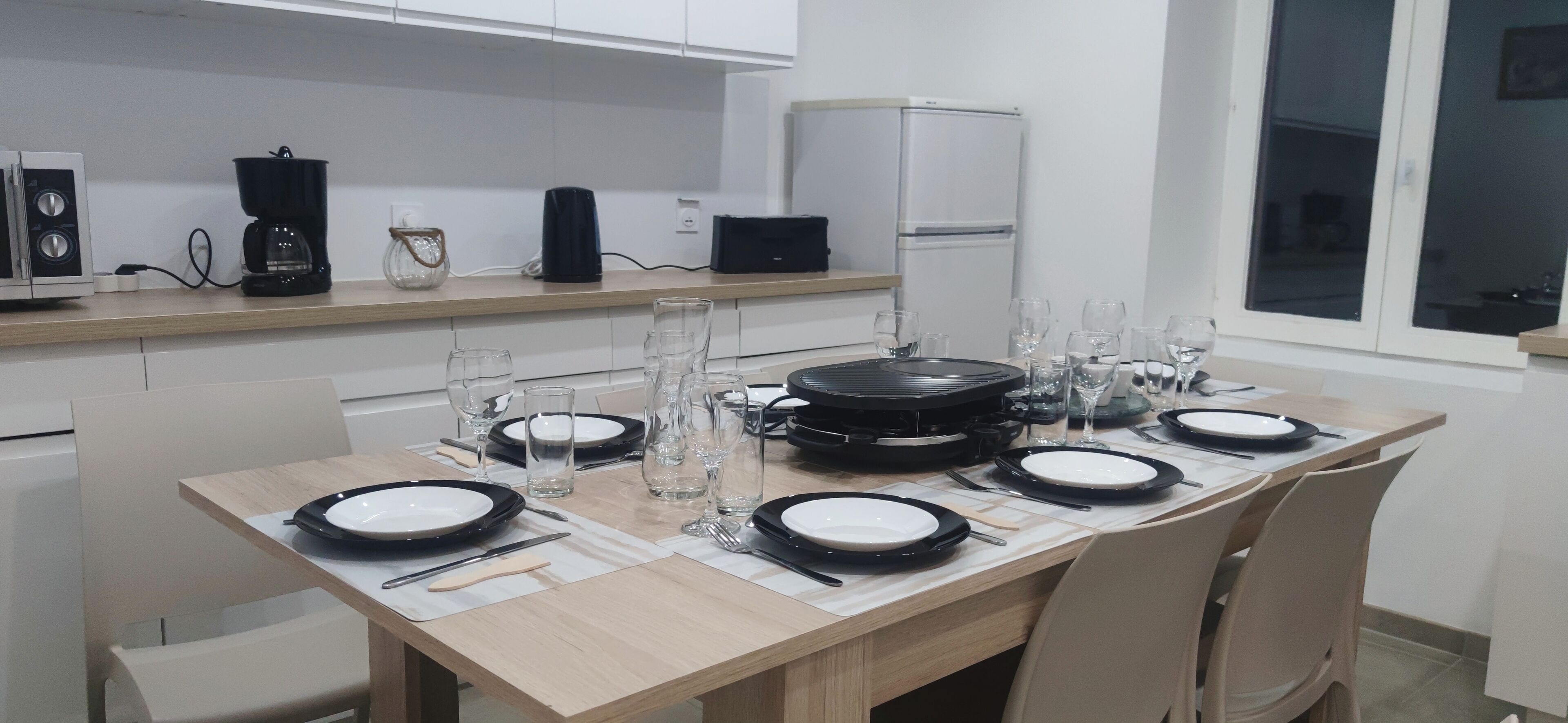 A modern kitchen with a dining table set for four, equipped with a coffee maker, a kettle, and a refrigerator.