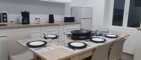 A modern kitchen with a dining table set for four, equipped with a coffee maker, a kettle, and a refrigerator.