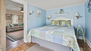 Un dormitorio con cama, mesita de noche, ventana con cortinas, ventilador de techo y un sofá en la habitación contigua.