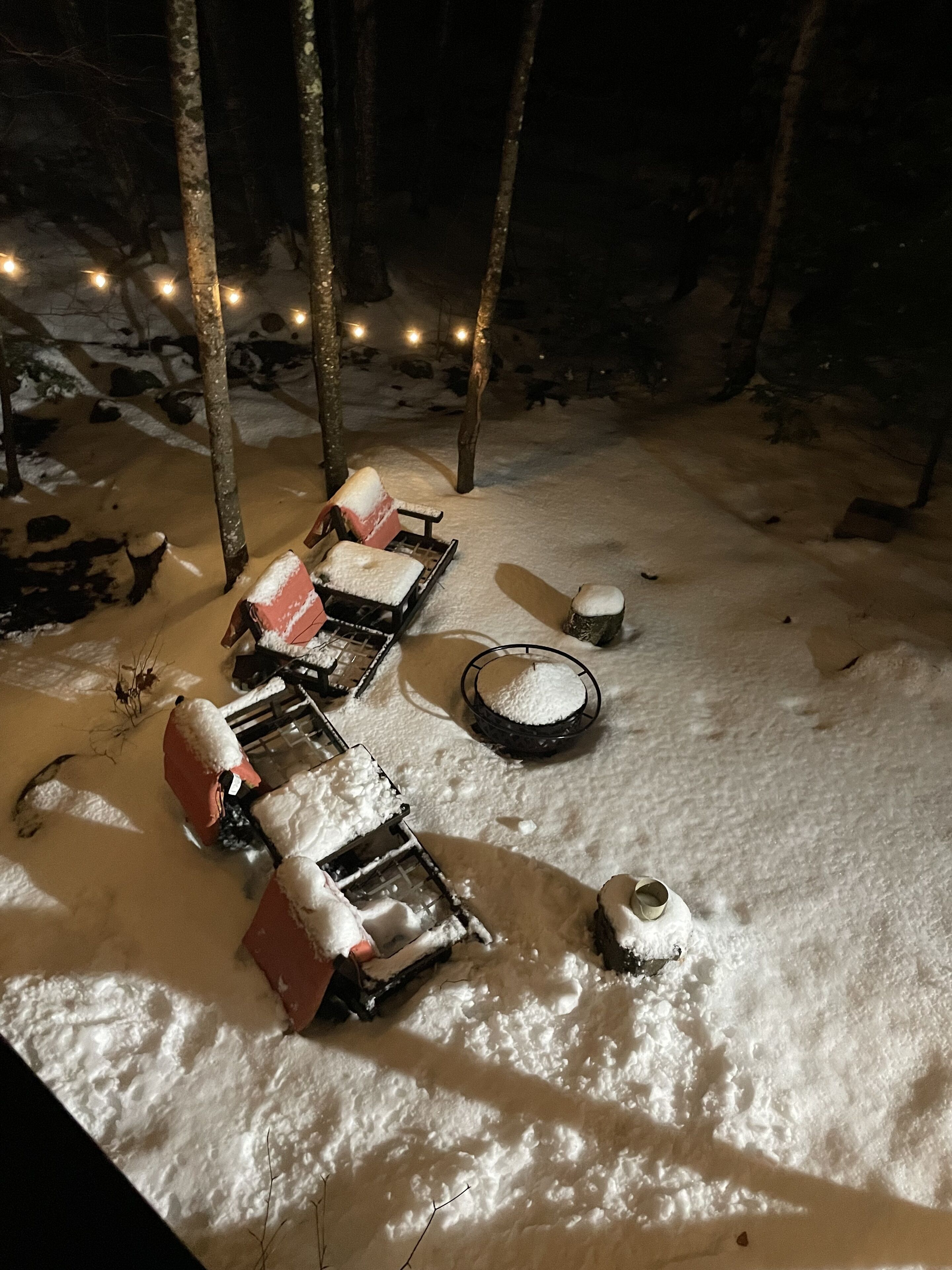 A snow-covered outdoor seating area with tables and chairs, surrounded by trees and illuminated by string lights.