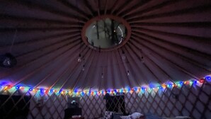 A yurt with a round central opening, colorful string lights, a small heater, and a bed with a blanket. - A yurt in the trees.  Close to Portland but close to nature.  Treeyurt Maine (Cumberland)