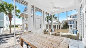 A spacious balcony with a wooden table and wicker furniture, large windows offering views of neighboring houses and palm trees. - Seas The Moments: Next to beach walkover, across the street from the pool! (Panama City Beach)