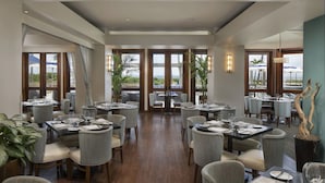 Un'ampia sala da pranzo con diversi tavoli e sedie, grandi finestre con tende e vista sul mare.