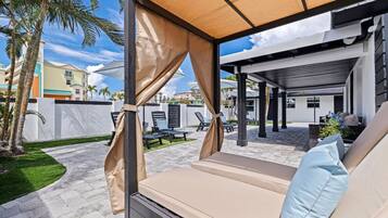 A covered outdoor seating area with a cushioned bench, a pergola structure, and a view of a residential neighborhood.
