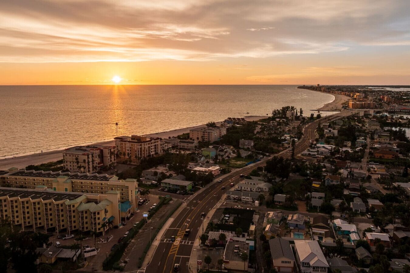 An aerial view of a coastal city with a beach, buildings, and roads during sunset.
