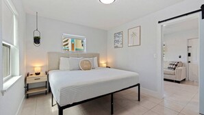 A bedroom with a bed, bedside tables, a hanging plant, a window, and a door leading to another room.