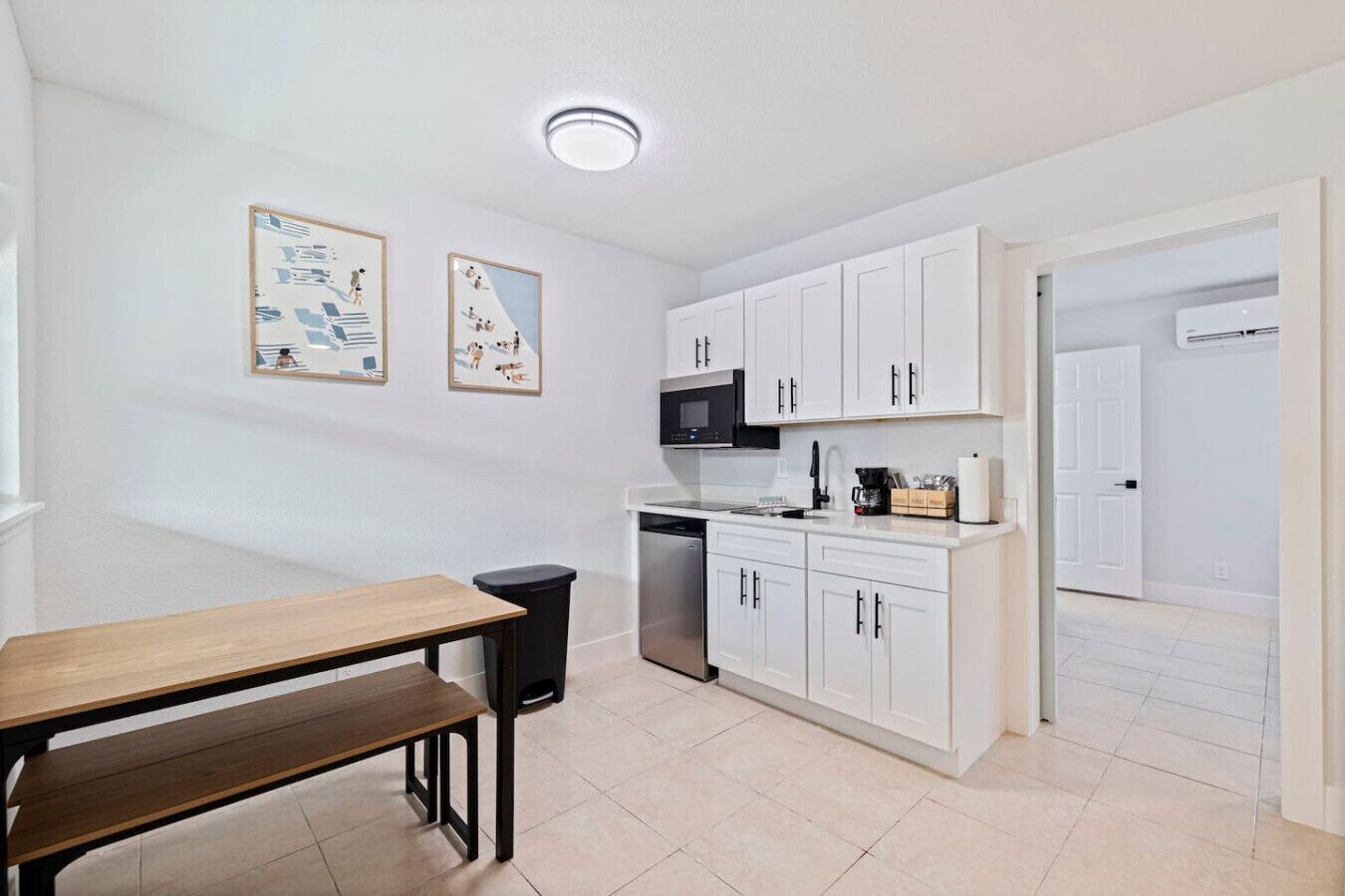 A modern kitchen with white cabinets, a black microwave, and a coffee maker. There is a wooden table and a trash bin.