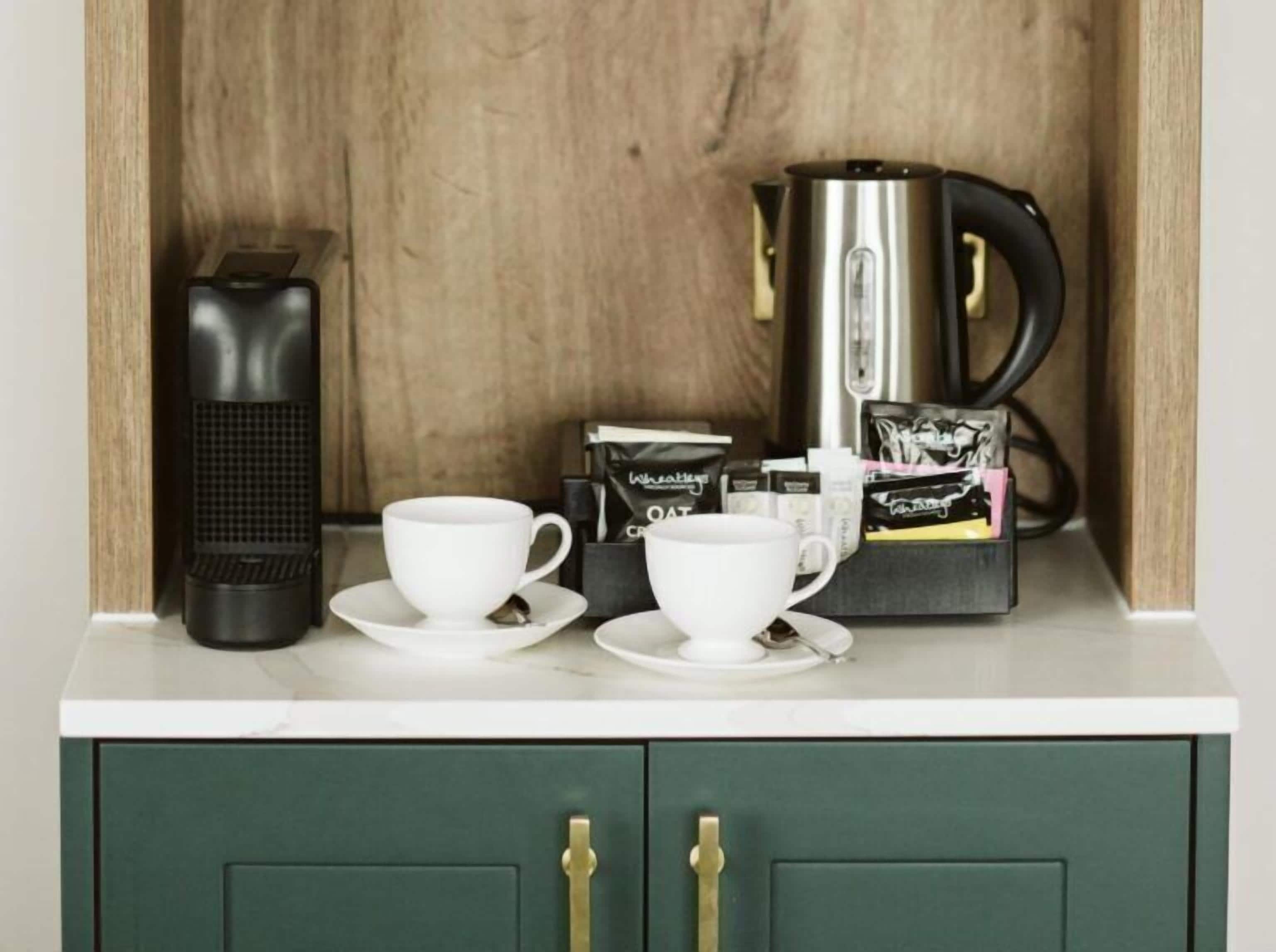 Una kitchenette con cafetera, dos tazas blancas, un infusor de té y cápsulas de café.
