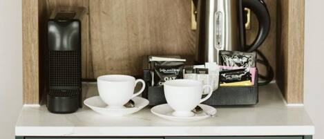 A kitchenette with a coffee maker, two white cups, a tea infuser, and coffee pods.
