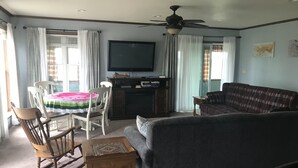 Een woonkamer met een bank, een houten schommelstoel, een tafel met een tafelkleed met watermeloenprint, een televisie en een plafondventilator.