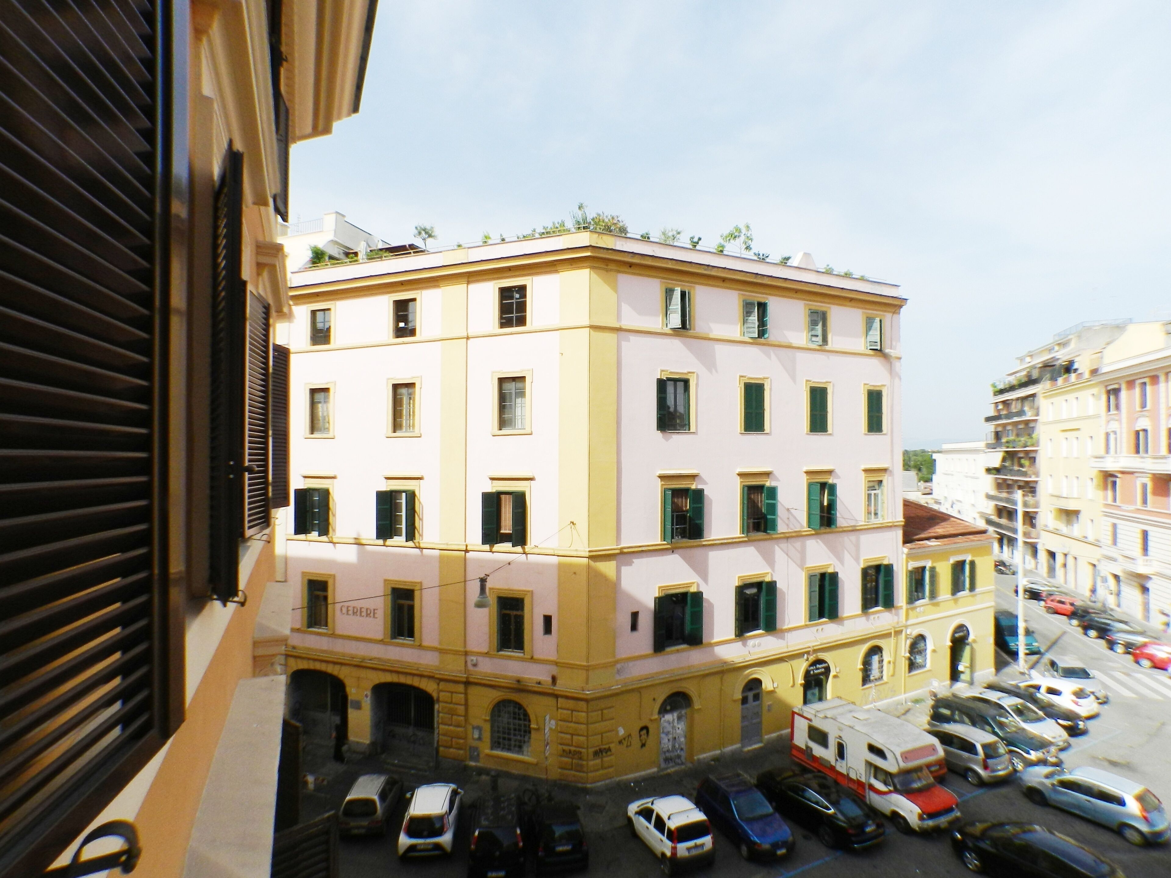 A multi-story building with a yellow facade and green shutters, featuring a ground-level entrance and several parked cars in front.