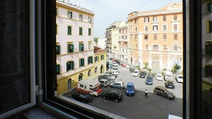 Blick aus einem Fenster auf eine Straße mit Gebäuden, parkenden Autos und einer Person, die unterwegs ist.