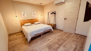 A bedroom with a wooden floor, a bed with a patterned bedspread, a bedside table, a hanging chair, and an air conditioning unit.