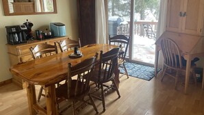A wooden dining table with chairs, a coffee maker, and a cabinet in a room with a large window and a door leading outside.