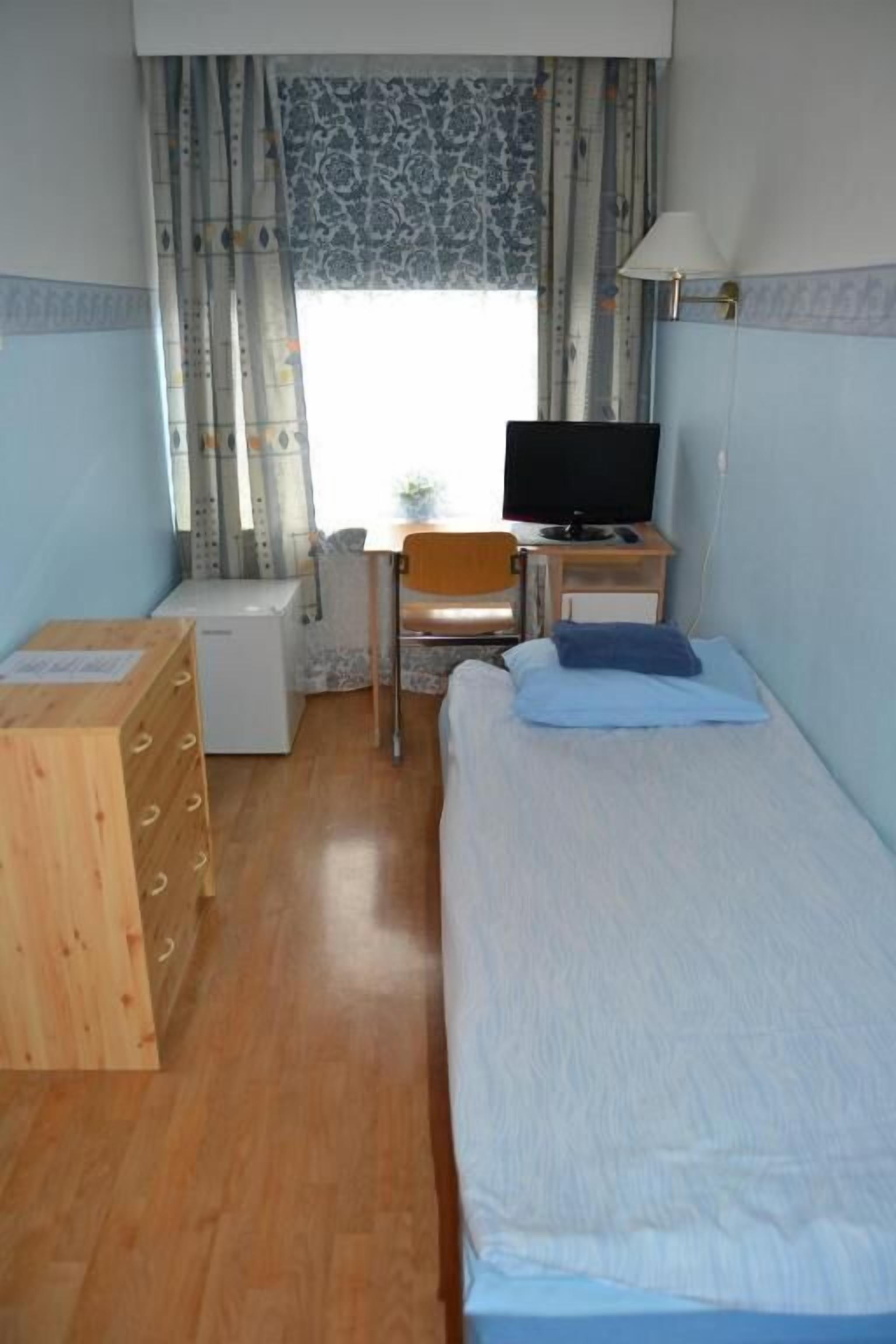 Ein kleines, ordentliches Zimmer mit einem Einzelbett, einer Holzkommode, einem Schreibtisch mit Stuhl, einem Fernseher und einem Fenster mit Vorhängen.