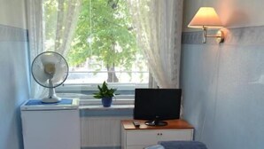 A compact room with a bed, a desk with a computer, a small radiator, a window with curtains, and a wall-mounted lamp. - Hostel Patria (Lahti)