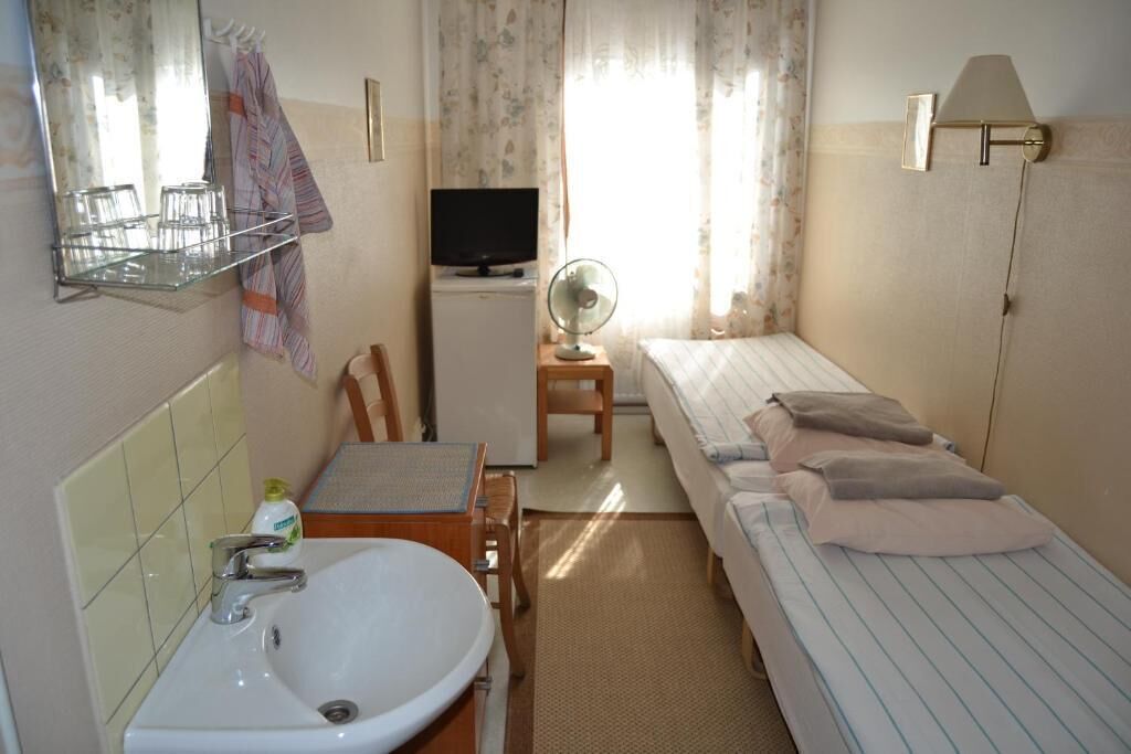 A small, well-lit room with a bed, a desk with a chair, a sink, a mirror, a television, and a fan.