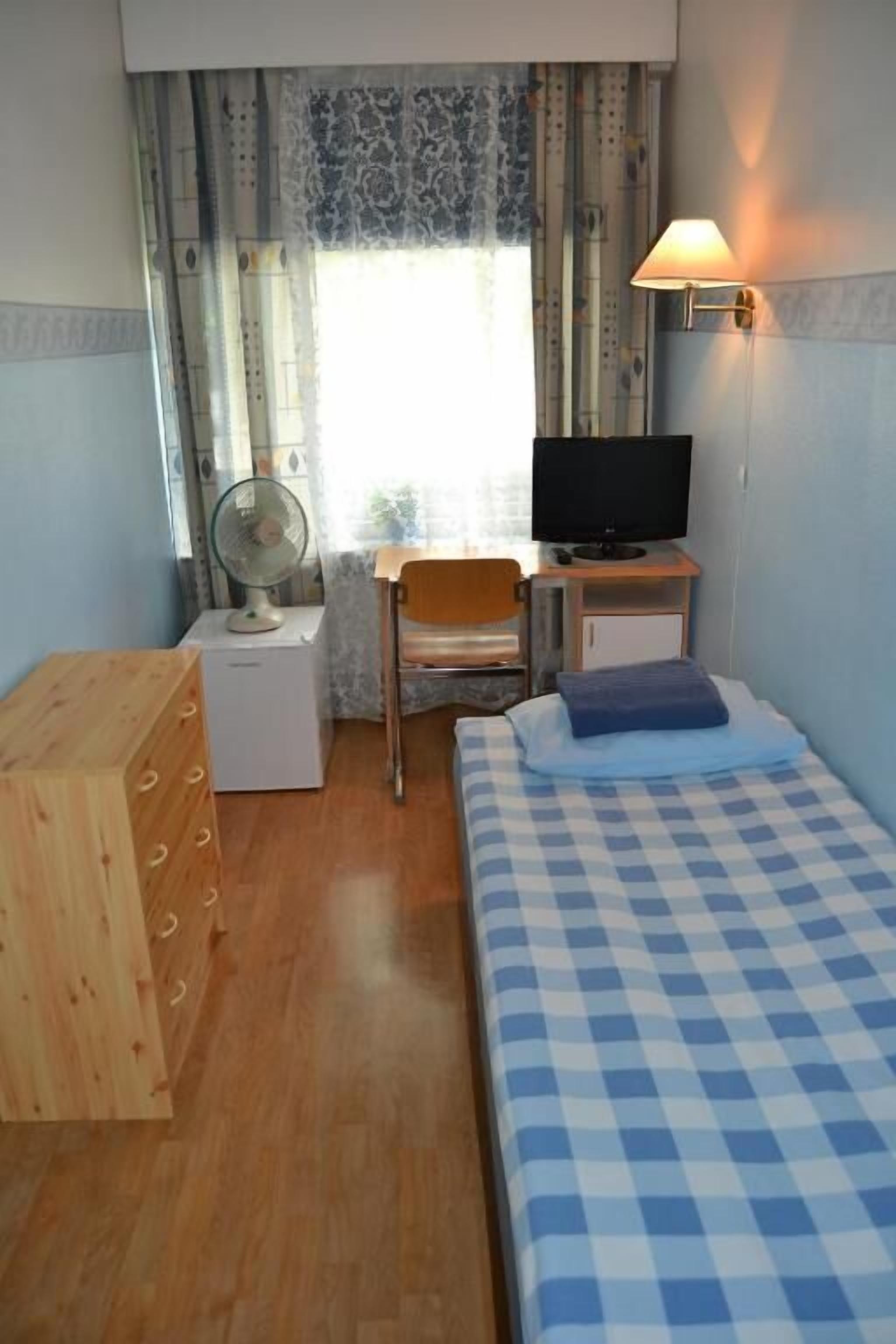 A compact room with a bed, a desk with a chair, a television, a fan, and a wooden dresser.