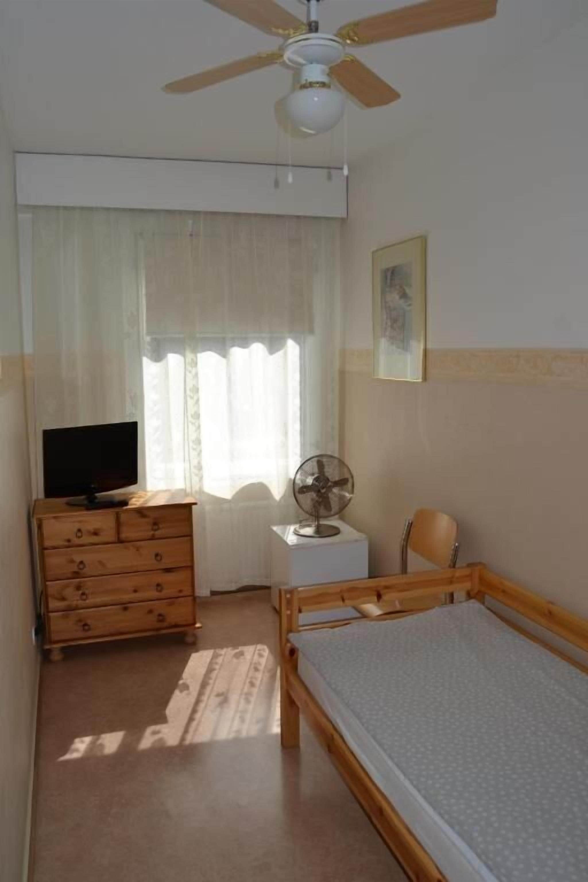 Une chambre à coucher avec un lit en bois, une commode, un téléviseur, un ventilateur de plafond et une fenêtre avec des rideaux.
