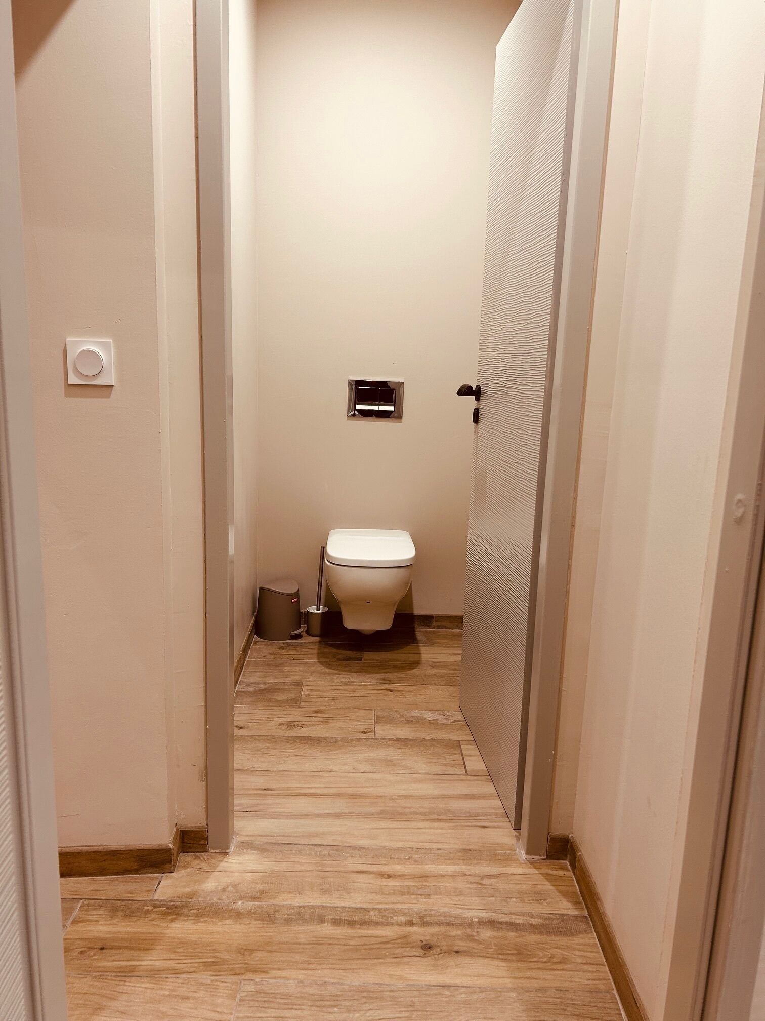 Un couloir étroit mène à une salle de bain équipée de toilettes, d’une petite grille d’aération et d’une poubelle.