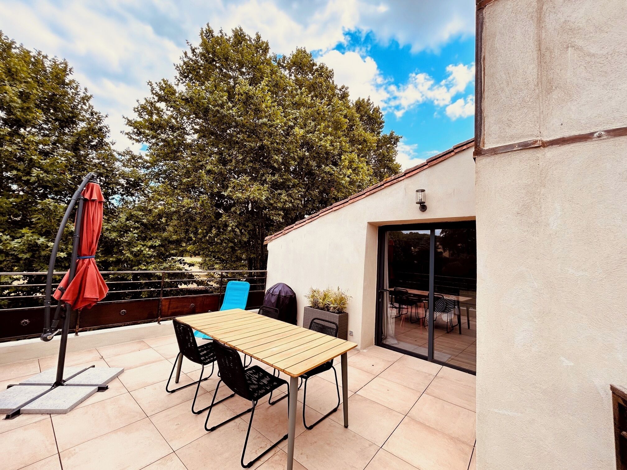 Un patio al aire libre con una mesa y sillas de madera, un paraguas rojo y un ventanal de vidrio que da a un espacio interior.