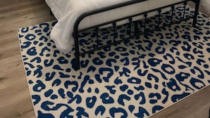 Ein ordentlich bezogenes Bett mit weißer Bettdecke und blauen Kissen, ein Leopardenfell-Teppich und Nachttischlampen auf kleinen Beistelltischen.