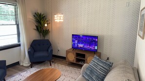 Ein Wohnzimmer mit einer Couch, einem blauen Sessel, einem Holztisch, einem Fernseher auf einem Holzschrank und einer Zimmerpflanze.