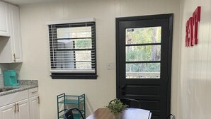 Eine kleine Küche mit weißen Schränken, einem Holztisch und schwarzen Stühlen. Es gibt ein Fenster mit Jalousien, eine Tür mit Aussicht und eine türkisfarbene Kaffeemaschine auf der Arbeitsfläche.