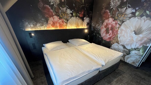 A neatly made bed with white linens in a room with a dark carpet and a floral mural on the wall. - Hotel Elsen (Grevenbroich)