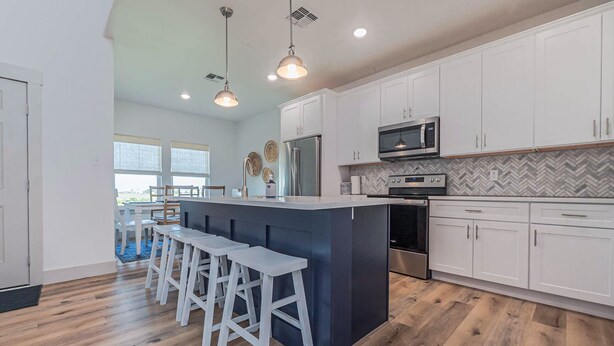 A modern kitchen with white cabinets, a central island with bar stools, stainless steel appliances, and a herringbone-patterned backsplash.