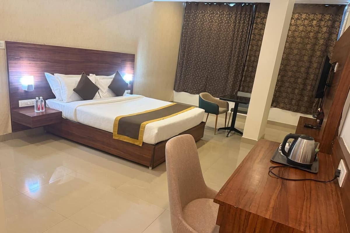 A hotel room with a bed, bedside table with bottles, a chair, a desk with a kettle, and curtains.