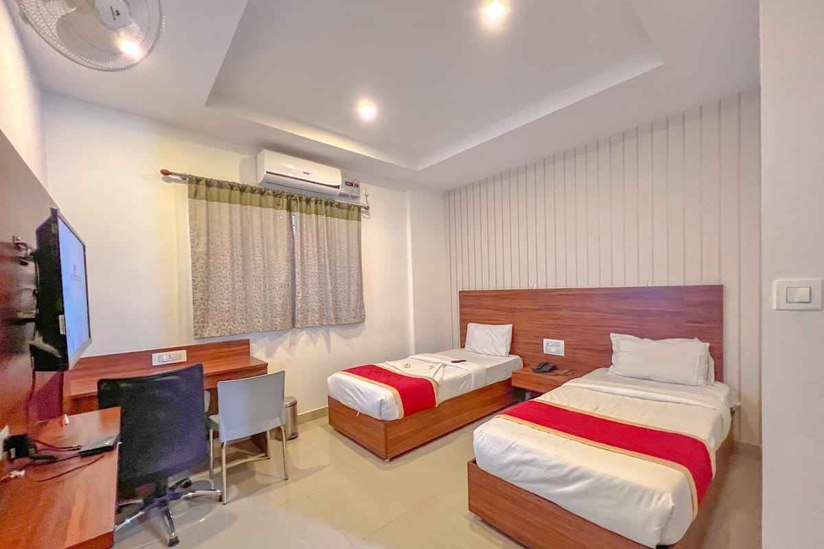 A hotel room with two single beds, a desk with a chair, a TV, and an air conditioning unit.