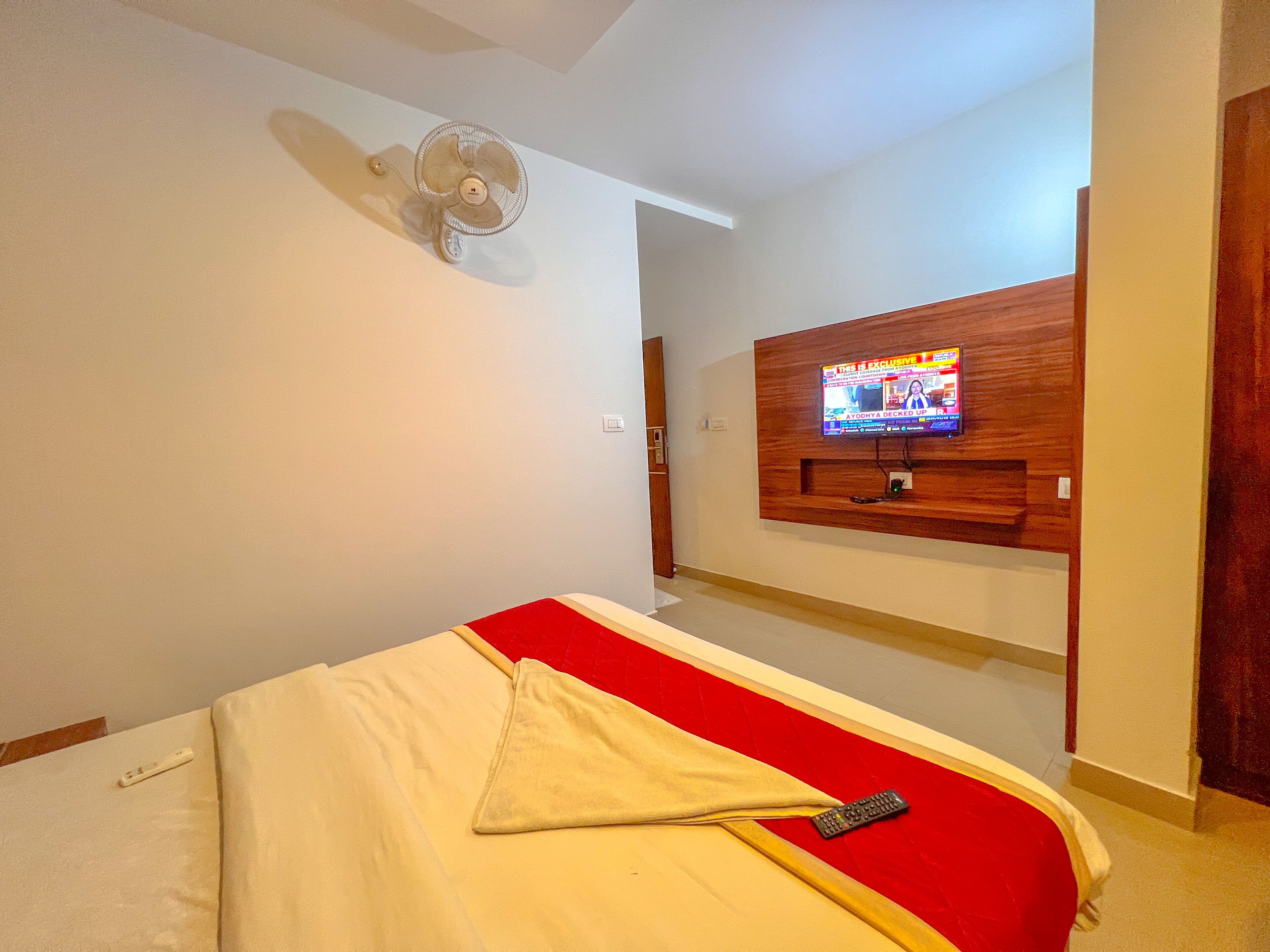 A hotel room with a bed, a TV mounted on the wall, a ceiling fan, and a red accent on the bed.