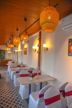 A restaurant with multiple tables, white chairs with red sashes, and hanging wicker lamps.