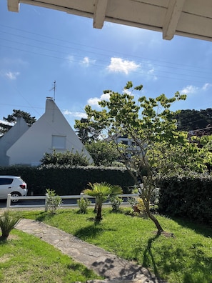 Een wit huis met een schuin dak, een witte auto geparkeerd ervoor en een verzorgde tuin met diverse planten en een grindpad.