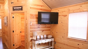 L'intérieur d'une cabane en bois, avec un téléviseur fixé au mur, une petite table agrémentée d'objets décoratifs et une porte donnant sur une autre pièce.