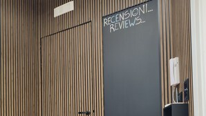 Une pièce avec un mur recouvert de lambris en bois, un tableau noir sur lequel on peut lire « RECENSI...REVIEWS... » et un panneau « BIENVENUE » posé sur le sol.