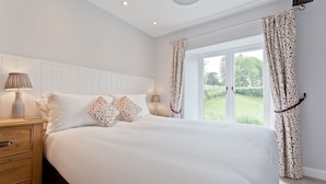 Un dormitorio con una cama grande, dos lámparas de noche, una mesita de noche de madera con cajones y una ventana con cortinas.