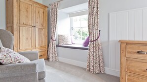 Una habitación con un armario de madera, un banco junto a la ventana con cojines morados y una cortina estampada.
